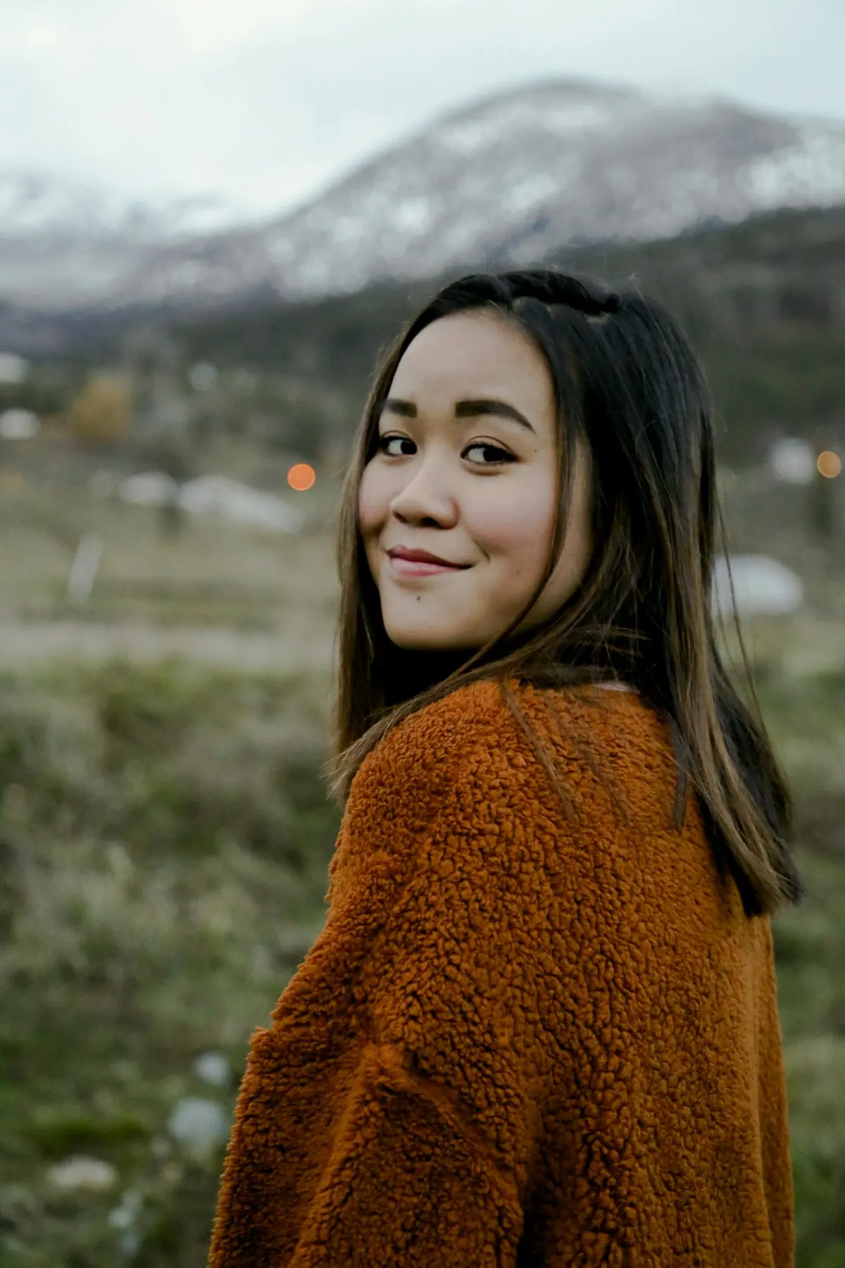 A woman in a rust-colored sweater looks back over her shoulder, mountains behind her.