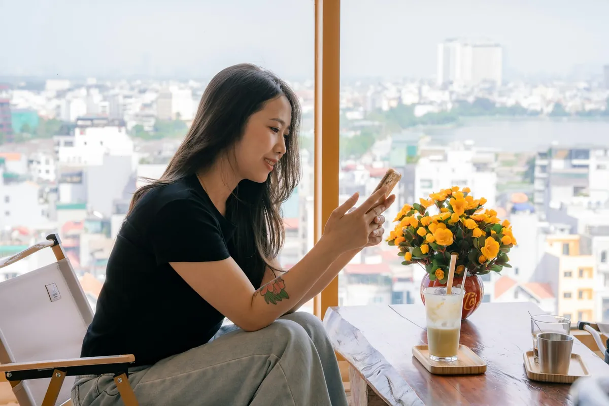 A woman sits by a wide window with her phone, the city beyond the glass.