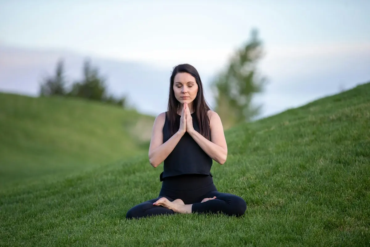 A woman sits in a meditation pose on a grassy hillside.